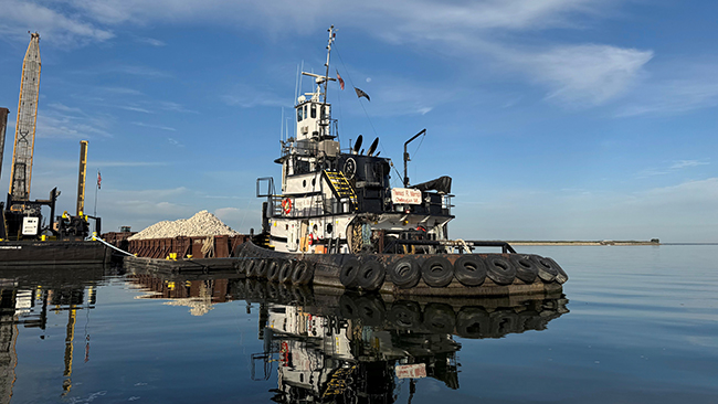 A photo taken from eye level of a tug pushing a barge with piles of cobble. The tug has tires lining the perimeter of its hull. The tug and barge are on water so smooth it looks like glass. Land is visible in the distance.